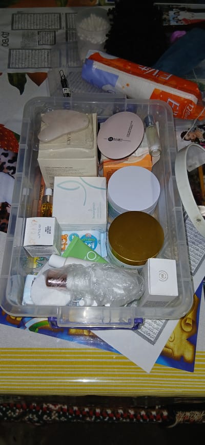 The image shows a transparent plastic storage bin filled with various beauty and skincare products. The bin is placed on a surface with a yellow and white striped pattern, which appears to be a table or mat. The contents of the bin include several boxes, bottles, and jars, suggesting a collection of personal care items. Among the visible items are a pink heart-shaped gua sha tool, a pink compact with "DR RASHEL" branding, a small clear bottle with a silver cap, a white jar with a gold lid, a box labeled "beautgreen," a green tube, and several smaller packages and bottles. Some items are wrapped in plastic. The background is cluttered with various items, including what appears to be a pack of sanitary pads, a cup filled with cotton swabs, and some papers with text. There is no indication of people or any specific location within Charshanba, Uzbekistan. The scene is a still life arrangement of personal cosmetic items.