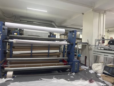 This image captures a busy industrial setting, likely a textile printing or processing facility, located in Bahçelievler, Turkey. The primary subject is a large industrial machine designed for handling rolls of fabric. The machine is blue and features several large rollers, some holding massive rolls of white and brown material. Some rolls appear to be decorative, with visible patterns. A smaller roll of white material is positioned at the bottom of the machine.
In the background, a man wearing a black t-shirt and what appears to be a black apron is visible, possibly a worker in the facility. He is standing near another piece of machinery, suggesting an ongoing operation. The setting is an indoor workshop, indicated by the concrete floor, exposed ventilation ducts, fluorescent lighting, and electrical conduits running along the ceiling and walls. The lighting suggests it could be daytime, but the specific time is not discernible. The overall atmosphere appears to be one of active production, with some material scattered on the floor, hinting at the dynamic nature of the work.
There are some visible labels and text on the machine, including a small screen displaying numerical data and a logo with the text "FM" and "TUAL MA." The scene conveys a sense of industry and functionality, with the focus on the large-scale textile processing equipment.