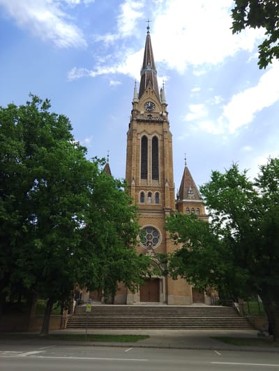 The image captures a majestic church with a tall, ornate steeple dominating the frame. The church is constructed of light brown brick, featuring arched windows and intricate stonework. A large circular stained-glass window is visible above the main entrance, flanked by wooden doors. Steps lead up to the entrance, with a sidewalk and a road in the foreground. Lush green trees frame the church on either side, their leaves providing a vibrant contrast to the architecture. The sky above is a bright blue, dotted with fluffy white clouds, suggesting a pleasant day. The sun appears to be high in the sky, indicating it's likely daytime.

A clock face is prominently displayed on the steeple, showing the time. There are no people visible in the image, and the scene appears serene and tranquil. The architecture suggests a European setting, possibly a historic town or city. A yellow sign with black text is visible near the steps, but the text is too blurry to read. The overall impression is one of grandeur and solemnity, with the church serving as a focal point amidst the natural beauty of the surrounding trees and the expansive sky. The presence of a road in the foreground hints at a public space.
