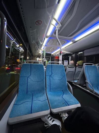 The image captures the interior of a modern public transport vehicle, likely a bus or tram, taken from a sideways perspective (rotated 90 degrees clockwise). The scene shows a brightly lit, clean, and futuristic-looking cabin, suggesting it is evening or night due to the reliance on artificial lighting and reflections from outside.
On the right side of the image, several rows of light blue upholstered seats are visible. These seats feature a subtle, darker blue geometric or abstract line pattern. The seat backs and bases are framed with light grey or white material. In the immediate foreground, a person's lower legs and feet are visible, wearing dark trousers and white Adidas sneakers with black stripes, resting near an empty seat. Further down the aisle, two other individuals are seated. One person, seen from behind, wears a green or olive-colored beanie/hat. Beside them, another person's head, possibly balding, is partially visible.
The left side of the image showcases the vehicle's interior wall and ceiling. Sleek, light grey or white panels line the area, illuminated by prominent horizontal LED strip lighting. These lights emit both cool white and blue hues, casting a distinct glow throughout the cabin. Above the seating area, a white, curved grab bar extends from the wall towards the center, ending in a handle with a red button or indicator. Higher up, another similar but shorter grab bar is present. The upper wall panels feature a perforated or mesh design.
Near the bottom left of the image, on a lower side panel, a "no smoking" symbol is clearly visible: a red circle with a diagonal line over a cigarette icon. The floor of the vehicle appears dark and highly reflective, mirroring the internal lights and showing blurred reflections of what might be external city lights or other vehicles passing by, reinforcing the impression of travel at night. The overall impression is one of modern design, efficiency, and cleanliness within an urban transport setting.