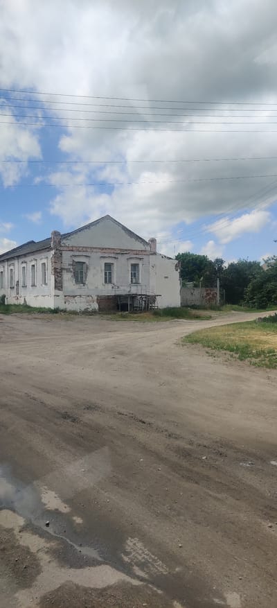 An old, two-story building with peeling white paint and exposed brick is visible on the left side of the image. The building has a gabled roof and several windows on the front facade. A small wooden porch with a railing is attached to the front of the building. In the foreground, a dirt road with puddles and tire tracks is visible. Trees and a metal fence are seen in the background. The sky is cloudy with some patches of blue. This scene is located in the city of Kolomak, Ukraine.
