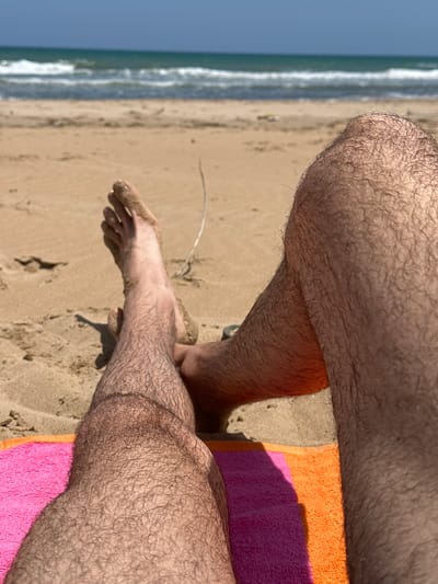 The image, captured from a first-person perspective in Guardamar del Segura, Spain, depicts a person's legs and feet resting on a striped beach towel. The towel features distinct pink and orange sections. The legs are covered in visible hair, and one foot is partially submerged in the light-brown sand of the beach, showing sand residue. The sandy foreground transitions into the mid-ground where gentle white-capped waves break in the turquoise sea. A clear blue sky is visible above the ocean horizon. Bright sunlight illuminates the scene, casting shadows.