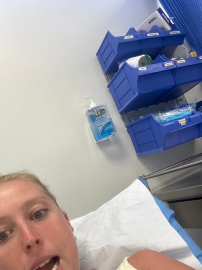The scene depicts a clinical or medical facility located in Corrimal, Australia. A female subject, with blonde hair, fair skin, and blue eyes, occupies the lower left portion of the frame. She lies on her back on a medical bed, which is covered with a white disposable paper sheet over a blue pad. Her mouth is open, and she looks directly towards the camera. The background features a plain, light-colored wall. Mounted on this wall is a 1-liter pump dispenser bottle of AQUIUM hand sanitizer. To the right of the hand sanitizer, a vertical system of blue plastic storage bins is mounted on the wall. These bins are labeled for medical supplies, with visible labels including "SWABS," "TISSUES," "CUPS," "GOWNS," and "MASKS." A white box with "winc." branding is situated on top of the uppermost blue bin. A blue privacy curtain is partially visible along the far right edge of the image.