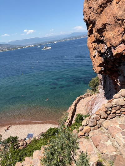This image captures a scenic view of the Esterel Massif coast near Théoule-sur-Mer, France. In the foreground, a steep, rocky cliff face dominates the right side of the frame, with a winding stone pathway descending along its edge, partially obscured by green foliage. A small, narrow beach made of pebbles is visible at the base of the cliff. The clear, turquoise waters of the Mediterranean Sea stretch out to the horizon, dotted with yellow buoys. In the distance, a coastline with buildings and hills forms the backdrop, with several yachts and boats sailing on the sea. Two individuals are swimming in the water, and two people are sunbathing on the beach. The sky is a bright blue with scattered white clouds, indicating a sunny day.
