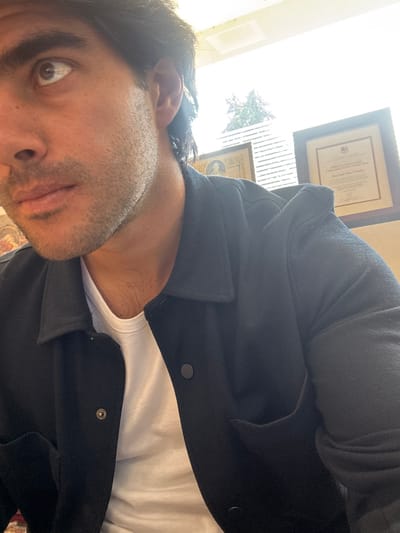 A man is shown from a low angle wearing a white t-shirt and a dark jacket with buttons. Part of his face is visible, showing a brown eye, his nose, and a portion of his mouth. Two framed documents are on the wall behind him and to his left. Light is coming in from a window, suggesting daytime. Part of a tree or bush is visible through the window.
