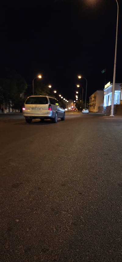 A silver station wagon is driving down a dark, paved street at night in Türkmenbaşy, Turkmenistan. Streetlights illuminate the road, and buildings can be seen on the right side of the street. A flag is flying on a pole in front of one of the buildings. The car's taillights are on, and its license plate is visible. The scene is quiet and appears to be a typical night on a city street.