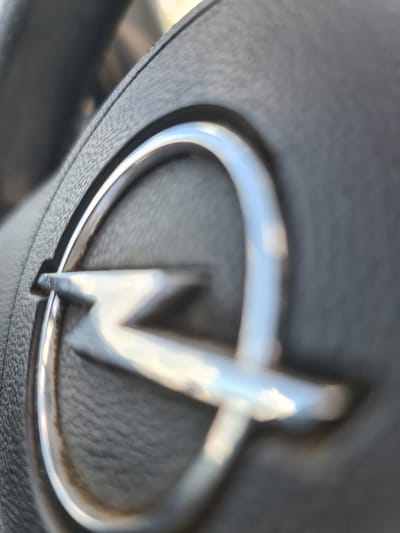 The image is a close-up, shallow depth of field photograph of an Opel car logo, likely on a steering wheel, as indicated by the textured black material surrounding it. The logo is a stylized silver lightning bolt within a circle. The background is blurred and dark. The photograph was taken in Maznovadnieki, Latvia, and depicts a car interior. There are no people or any action visible in the image.