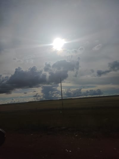 This is a landscape photograph taken in Қонысбай, Kazakhstan. The image captures a vast, open field under a dramatic sky. The sun is visible in the upper part of the frame, partially obscured by thick, dark clouds, creating a somewhat overcast and possibly stormy weather condition. Rays of sunlight pierce through the clouds, illuminating parts of the sky and casting a soft glow on the scene.
In the foreground, a dark, unpaved road or ground is visible, with some scattered debris. A single, tall wooden pole stands upright in the middle ground, likely a utility pole, with faint power lines stretching across the sky. Beyond the pole, a wide expanse of golden-brown grassland stretches towards the horizon, suggesting it might be a dry field or agricultural land. The horizon is defined by a slightly elevated terrain, dotted with distant structures or formations.
The overall mood of the image is somewhat somber due to the heavy clouds and the limited visibility of the sun, but the light breaking through offers a touch of hope or drama. The time of day appears to be late afternoon or early evening, given the angle of the sun and the quality of light. There is no visible text in the image.