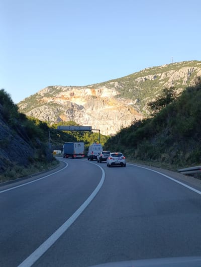 The image captures a view of a highway in Đurmani, Montenegro, under a clear blue sky. The road curves to the right, with a white line marking the lane. On the left side of the road, a steep, rocky slope covered in vegetation rises sharply. On the right, the road is bordered by a guardrail and more dense vegetation.
In the background, a large, excavated mountainside with exposed rock formations dominates the landscape. The rock exhibits shades of orange, beige, and white, suggesting it is a quarry. Green trees and shrubs cover the upper parts of the mountains and the surrounding slopes.
Several vehicles are visible on the highway. A large blue semi-trailer truck is in the left lane, followed by a white van. To the right of the van, a white hatchback car is in the right lane. A large overhead sign structure spans the road in the middle distance.
The lighting suggests it is either early morning or late afternoon, with long shadows cast by the vehicles and the terrain. The overall scene depicts a rural or semi-rural highway passing through a mountainous area.
