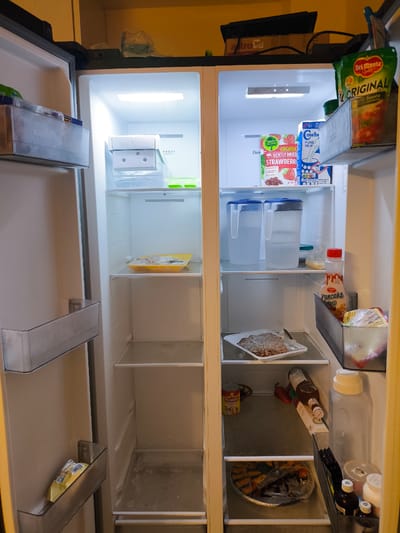 The image shows the inside of an open refrigerator, likely in a home kitchen in Imus, Philippines. The refrigerator has two vertical doors, one on the left and one on the right, and is illuminated by interior lights.

The left side of the refrigerator contains several shelves and drawers. The top drawer is partially visible and contains some items. Below that, a clear shelf holds a yellow plastic tray with some food items on it. The lower shelves are empty. The door of the left compartment has a transparent bin attached to it.

The right side of the refrigerator is more densely stocked. The top shelf has a bag of Del Monte Original Style Tomato Sauce, a box of "Cowhead" Pure Milk, and a bag of "Berry Village" Organic Gently Dried Strawberries. Below that, two large clear pitchers filled with water are visible, along with a small green container. Further down, a bottle of pancake syrup is prominent, next to a container of what appears to be ground meat or a patty in a white plastic tray. The next shelf down holds a can of "Alaska" Evaporated Milk and a red chili pepper. The bottom shelf contains a round aluminum tray filled with food, possibly pastries or appetizers, covered in plastic wrap. The door of the right compartment has several small containers and bottles, including some with dark liquid.

The overall impression is that of a moderately stocked refrigerator. There are no people visible in the image. The lighting suggests it could be any time of day.