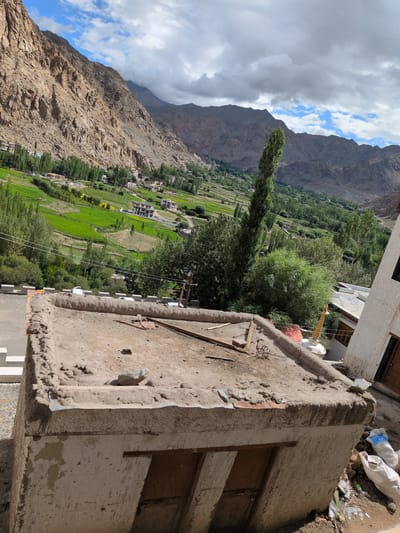 The image shows a view of a rural village nestled in a valley, with rugged, brown mountains dominating the background. In the foreground, there is a rough, concrete structure with a flat, debris-strewn roof. The roof has some wooden planks and scattered stones on it. Adjacent to this structure, a white building with a brown door is visible on the right.

Below the foreground structure, the landscape opens up to a vibrant green valley with terraced fields. Numerous trees are scattered throughout the valley, and several houses with white walls and flat roofs are visible, indicating a settlement. A tall, slender tree stands out in the mid-ground. The sky is partly cloudy with patches of blue sky peeking through.

The scene depicts a mountainous region, likely in Ladakh, India, given the architecture and landscape. There are no people visible in the image. The overall impression is one of a remote, agricultural community surrounded by dramatic natural scenery.