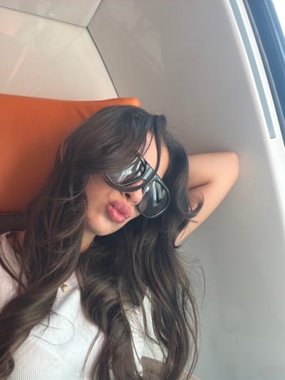 A young woman with long, dark, wavy hair is captured in a close-up, tilted shot, lying down in what appears to be a modern, light-colored interior, likely a vehicle or boat cabin in Saint-Laurent-du-Var, France. She is wearing large, black sunglasses that obscure her eyes, and her lips are pursed in a slight "kissing" expression, accented by pink lipstick. Her head rests comfortably on an orange-colored cushion or headrest. One arm is bent and raised, with her hand positioned behind her head. She is dressed in a light-colored top, and a small, gold-colored charm is visible near her chest, partially covered by her hair. The background features smooth, white or off-white panels and lines, suggesting structural elements or trim, with a darker and lighter strip overhead that resembles a window frame or sunroof. The lighting is bright and even, indicating it is daytime. The image conveys a sense of leisure and a relaxed pose for a photograph.