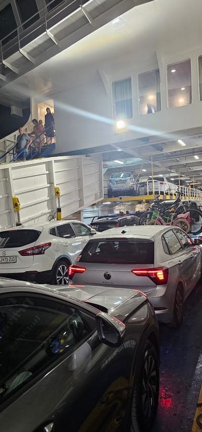 The image captures the interior of a ferry, likely in the city of Supetar, Croatia, where vehicles are parked on multiple levels. In the foreground, several cars are visible, including a silver Volkswagen Polo with its taillights illuminated, and a white Nissan Qashqai with a license plate reading "SJ 73-ZUD". To the left, a dark-colored car is partially visible, reflecting its surroundings. In the background, a silver car is parked on an upper level of the ferry, accessible by a ramp. Several bicycles are also parked on the ferry. On the upper deck, a group of people are seen walking down a staircase. The environment is industrial, with exposed metal structures and lighting fixtures.