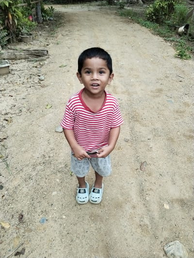 The image depicts a young child standing on a dirt path in Teupinbayu, Indonesia. The child, appearing to be male, has dark hair and a dark complexion, estimated to be between 2 and 5 years old. He is wearing a red and white horizontally striped short-sleeved t-shirt, light grey shorts patterned with repeating cartoon rabbits, and light blue or grey closed-toe sandals featuring a black and white cartoon face design with eyes and a smile on the front. The child is positioned centrally, facing directly towards the camera with his mouth slightly open, suggesting a smile or verbalization. Both hands are clasped in front of his waist, holding a small, flat, dark-colored object, possibly a mobile phone or toy.

The immediate environment is an unpaved, light brown dirt path, characterized by loose soil, small pebbles, scattered dry leaves, and sparse green vegetation. The path extends into the background. To the left of the path, dense tropical foliage, including large green leaves, tree trunks, and several unglazed terracotta-colored pots, is visible. A weathered wooden log or branch lies horizontally on the ground near the left edge of the path. To the right of the path, there is additional greenery, with some plants and another container or object partially visible in the mid-distance. The scene is illuminated by natural daylight, indicating an outdoor setting.