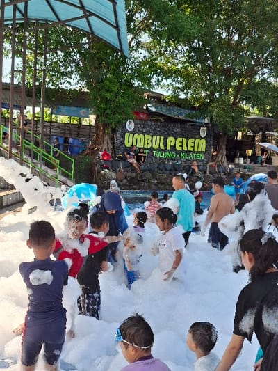 A group of people are playing in a foam pit. They are located at Umbul Pelem, Tulung, Klaten, Indonesia. There is a large sign above the foam pit that reads "Umbul Pelem Tulung Klaten". The people are mostly children and they are all having fun playing in the foam. There are many adults watching them, some are standing on the edge of the pit while others are seated on the ground. The environment is mostly green with trees and lush foliage in the background. The foam pit is surrounded by a rock wall. The sky is blue and there are a few clouds. The people are dressed in casual clothes, with some wearing swimwear. There is a large white canopy in the background, providing shade from the sun. Some of the children are splashing each other with foam while others are jumping and laughing. There is a blue inflatable ring floating in the foam pit, adding to the fun atmosphere. The scene is captured from a slightly elevated perspective, offering a clear view of the action in the foam pit.