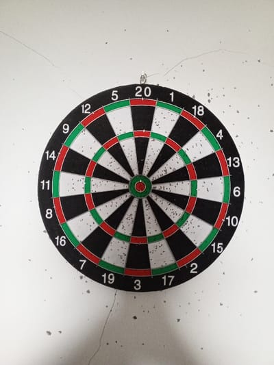 A standard dartboard is centrally mounted on a textured, off-white wall in Al Haouafate, Morocco. The circular dartboard features traditional black and white segments, with red and green rings for double and triple scores, and a red bullseye surrounded by a green ring. The numbers 1 through 20 are printed in white around the perimeter. Numerous small, dark impact marks are visible across the dartboard's surface, particularly within the white segments, indicating prior use. The dartboard is suspended by a single, small metallic hook protruding from the wall above the "20" segment. The wall itself exhibits several fine, linear cracks, notably a curved one originating from the upper-left quadrant and extending towards the center-top. Scattered dark specks, possibly debris or imperfections, are present across the wall's surface. No individuals, actions, or other objects are depicted.
