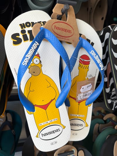 The image displays a close-up of several pairs of Havaianas flip-flops, featuring a design of Homer Simpson. The flip-flops are white with blue straps and depict Homer in his signature yellow skin and red briefs. One pair is prominently displayed in the foreground, with its sole showing Homer's full body and the brand name. A price tag is attached to this pair, indicating €29.99. Another pair, also with Homer Simpson, is partially visible to the right. In the background, other Havaianas flip-flops and footwear are on display, including some with black and white patterns. The setting appears to be a retail environment, possibly a store in Saint-Raphaël, France, given the context provided. The overall lighting suggests an indoor setting, possibly a shop.
