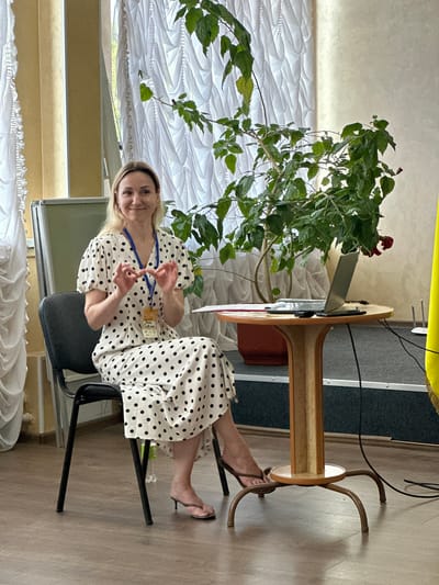 A woman with blonde hair is sitting in a black chair in what appears to be a conference room or lecture hall in Myronivka, Ukraine. She is wearing a white dress with black polka dots and has a lanyard around her neck with a badge. The woman is gesturing with her hands as if speaking. In front of her is a round wooden table with a laptop, a phone, and some papers. A large potted plant with green leaves is positioned behind the table. The room has white curtains in the background and a yellow flag on the right side. The floor is made of dark grey carpet and wooden planks.