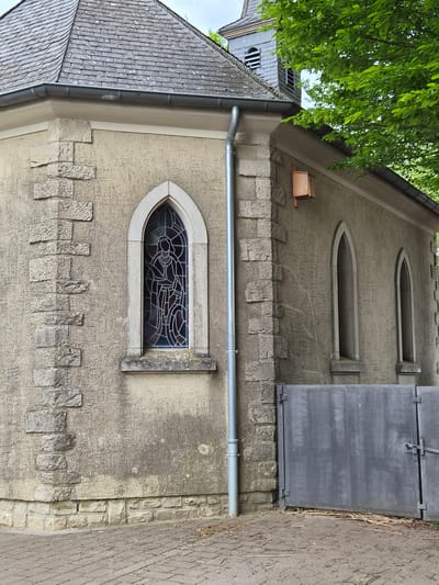 The image depicts the exterior of a building, potentially a church or chapel, situated in Schengen, Luxembourg. The building is constructed with textured, light-colored walls. A distinctive feature is a stained glass window with an arched top. Above the window, there are two smaller, horizontal slit windows. A gray metal door or covering is present to the right of the upper windows. A gray gutter runs horizontally along the wall above the stained glass window. The building's roof is made of gray tiles and has a drainpipe. Green foliage is visible in the upper left corner of the image. The side of the building is flanked by a stone paved walkway. The image is taken at an oblique angle.