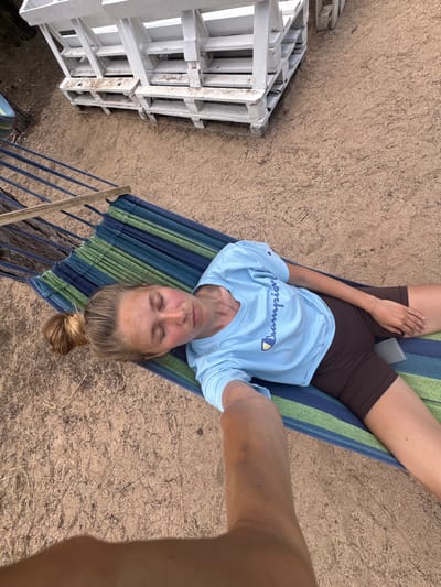 A woman is lying in a hammock in a sandy outdoor area, possibly a beach or campground. She is wearing a light blue Champion t-shirt and brown biker shorts. Her eyes are closed, suggesting she is resting or sleeping. The hammock is striped in shades of blue and green. In the background, there are white wooden structures, possibly benches or tables, and some dry vegetation on the sand. A person's arm is visible in the foreground, holding a phone to take the selfie. The overall impression is one of relaxation in a rustic outdoor setting.