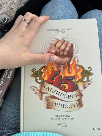 The image presents a close-up, overhead view of an individual's lap, located in the city of Yus'ki, Russia. A light-skinned left hand, wearing a thin silver-colored ring on the ring finger and exhibiting clear-to-light-pink ombré manicured nails, is positioned on the right side of a cream-colored hardcover book. The book's cover features Russian text, including "МАКСИМ БАТЫРЕВ (КОМБАТ)" at the top, and "45 ТАТУИРОВОК ЛИЧНОСТИ" with "ПРАВИЛА МОЕЙ ЖИЗНИ" at the bottom, along with a "МИФ" publisher logo. The central illustration on the cover depicts a stylized clenched fist emanating light rays, situated above the large red numbers "45" surrounded by orange flames, all overlaid on a beige banner. The book rests upon blue denim jeans. To the left, partially obscured, is a second book with a dark brown spine, visible with the text "НАКОПИТЕЛЬНЫЙ ЭФФЕКТ". In the upper left corner, a dark fabric with a silver-colored zipper pull is visible. A light blue, ribbed knitted fabric is partially discernible at the bottom edge of the frame.