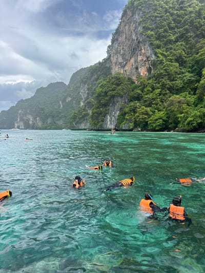 A group of people are snorkeling in the clear turquoise waters off the coast of Haad Hin Khom, Thailand. The jungle-covered limestone cliffs rise steeply from the water in the background, creating a dramatic natural backdrop. The sky is overcast with dramatic clouds, suggesting a recent or impending rain shower. Several boats are anchored in the distance, and a few individuals are visible on paddleboards. The snorkelers, all wearing orange life vests and snorkeling gear, are spread out across the frame, enjoying the underwater scenery.