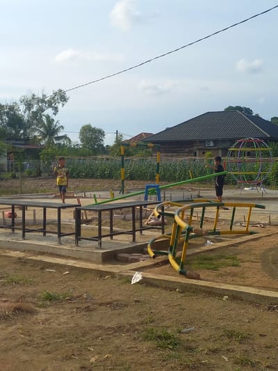 The image depicts an outdoor playground area under a clear sky with scattered clouds, likely located in Buketseuraja, Indonesia, given the tropical foliage and architectural style of the buildings visible in the background. In the foreground, a partially constructed playground features concrete foundations and metal structures for various play equipment. A yellow and green seesaw is positioned to the left, supported by a blue base. To the right, a large, colorful spherical climbing frame is visible, constructed from metal tubes.
Two young boys are present. The boy on the left, wearing a yellow t-shirt with a graphic print and dark shorts, is standing on a concrete platform. The boy on the right, wearing a black t-shirt and black shorts, is interacting with the green beam of the seesaw, pointing towards it. The ground surrounding the playground is a mix of dirt and sparse grass, with some construction debris scattered about, suggesting the playground is newly built or undergoing renovation. In the background, residential buildings with dark tiled roofs are nestled amongst lush green vegetation, including palm trees, and what appears to be cultivated fields. A power line stretches across the upper portion of the frame.