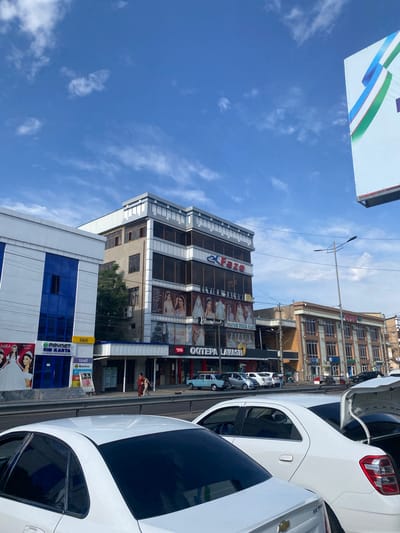 The image depicts a street scene in Parkent, Uzbekistan, under a bright blue sky with scattered white clouds. In the foreground, several white cars are parked or driving on a road. One white car in the immediate foreground has its trunk open.
Behind the cars, a row of buildings lines the street. The buildings vary in architectural style and age. On the left, a modern building with blue and white facades stands out. Further down the street, a multi-story building with glass windows displays advertisements and signage, including text that appears to be in a Cyrillic-based alphabet. One prominent sign features the word "Fazo" in large red letters. Another building showcases images of wedding dresses.
Further down, another building has a facade with numerous windows. A street light stands tall on the right side of the image. People are visible walking on the sidewalk in front of the buildings. The overall impression is a typical urban environment with commercial establishments and vehicular traffic.