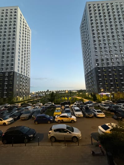 The image depicts a view of a residential complex in Drozhzhino, Russia. Two tall, modern apartment buildings, characterized by their numerous windows and balconies, dominate the left and right sides of the frame. Between the buildings, a large parking lot is filled with a variety of cars, including sedans, SUVs, and what appear to be taxis. The sky above is a clear blue, transitioning to a lighter shade near the horizon, suggesting it is either early morning or late evening. In the background, more buildings and infrastructure, possibly another part of the residential complex or surrounding urban development, are visible, illuminated by streetlights. The foreground shows a paved walkway with a few vertical bollards, and a dark-colored car parked on the left. A dark-colored electric scooter is parked near some greenery on the right. The overall scene suggests a typical urban residential environment.