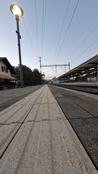 An outdoor, low-angle, eye-level shot captures a train station platform at dusk in Unknown, Unknown. The foreground features a tactile paving strip of light-colored, textured concrete blocks that leads into the distance. To the right of this strip are darker, grey concrete blocks. Beyond the platform, train tracks are visible, and a covered platform structure with lighting extends into the background. Overhead, a network of power lines crisscrosses the pale sky, supported by poles. On the left side of the image, a tall lamppost with an illuminated fixture stands next to a section of a house with a brown roof and a small awning. Green foliage borders the left side of the platform. The overall scene is devoid of people or activity, suggesting an empty or disused station. The lighting indicates it is either dawn or dusk.