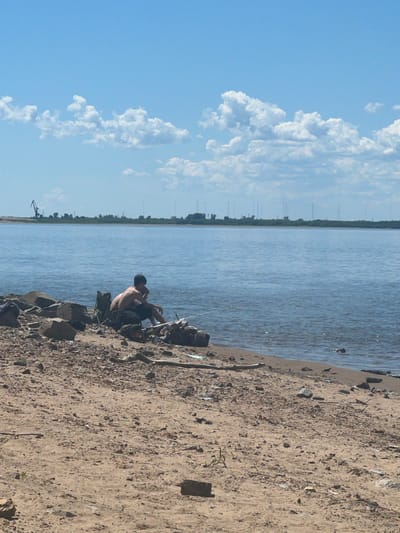 A person is fishing from the bank of a wide body of water. The person is sitting on a foldable chair on a rocky shoreline, facing the water. Fishing rods are placed in the water. The shoreline is sandy and littered with rocks and debris. In the background, across the water, is a treeline with various structures, including what appears to be a large crane on the left side. The sky is blue with scattered white clouds. The location is not identifiable as being in the city of имени Тельмана, Russia, as no specific landmarks or context confirm this.