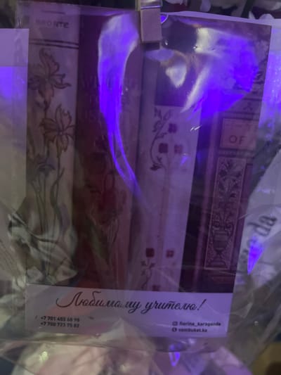 The image shows a close-up of several books, possibly a gift set, wrapped in clear plastic. The books have decorative covers with floral patterns and what appear to be embossed titles. One visible title fragment reads "BRONTE". There is also a label with Russian text that translates to "To my beloved teacher!" and includes contact information: phone numbers starting with "+7 701" and "+7 700", an Instagram handle "@florina_karaganda", and a website "vambuket.kz". The lighting in the image is predominantly purple, suggesting artificial light, and there are some reflections on the plastic wrap. The overall scene is dimly lit and focuses on the product itself, hinting at a commercial or personal gift-giving context. The location context provided is Shahtinsk, Kazakhstan.
