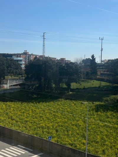 An elevated perspective reveals an urban landscape. A vibrant green field occupies the foreground, bordered by a concrete structure. Beyond, a tree-lined area transitions into residential buildings. Power lines stretch across a clear blue sky. A small, rectangular sign is visible at the edge of the green space. A zebra crossing is present on the concrete structure.