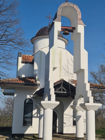 The image depicts the church "St. Nicholas the Wonderworker" in Shumentsi, Bulgaria. The church features white walls, a copper dome topped with a cross, and a bell tower with a bell. The architecture includes a series of arched openings and columns. The scene is captured on a bright day with a clear blue sky and bare trees in the background.