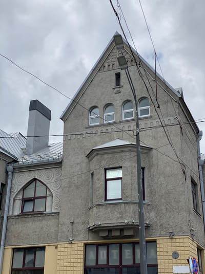 The image shows a cloudy day in Sortavala, Russia, with a focus on an old building. The building is constructed of grey stucco and features various architectural details. On the left, a prominent grey chimney with a dark grey cap rises from a metal roof. Below this, a decorative arched window with a dark frame is visible, surrounded by a stucco pattern of concentric circles. The main facade of the building has a distinct gable roof. Several arched windows with white frames are situated on the upper part of this facade, with a smaller rectangular window above them. A projecting bay window with a dark framed window is also present. Below the bay window, a narrow ledge is supported by decorative brackets. The lower part of the building is painted a light yellow and features a row of windows with dark frames. One of these windows has text that appears to read "АПТЕКА", indicating it is likely a pharmacy. The building is also adorned with decorative stucco detailing above the windows and along the roofline. A tall grey lamppost with two lamps stands in front of the building, partially obscuring the view. Numerous electrical wires crisscross the scene, indicating an overhead network. A corner of the building at the bottom right is visible, with a yellow tiled section and a traffic sign indicating restrictions. The overall impression is of a historic building in a somewhat overcast urban environment.