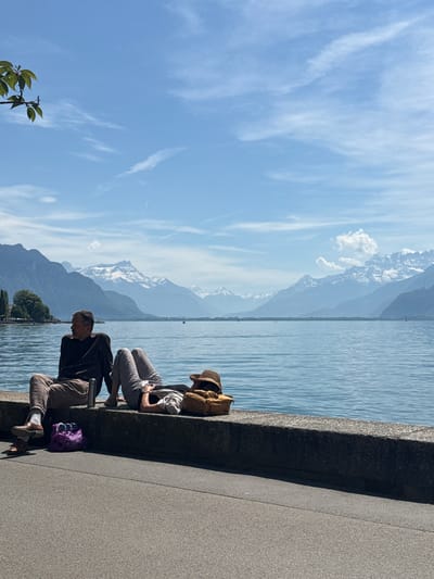 Two individuals are sitting on a stone wall overlooking a body of water, likely Lake Geneva, with a backdrop of mountains, some snow-capped. The setting appears to be in Vevey, Switzerland, given the lake and mountain scenery. One person is wearing a hat and has a backpack, while the other is wearing casual clothing. A purple bag and a metal object are near the second person. The sky is blue with scattered clouds.