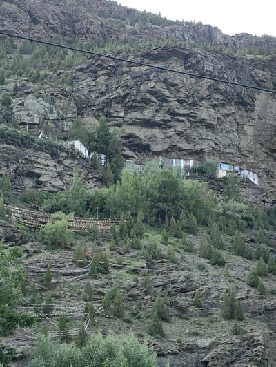 The image depicts a mountainous landscape in Kardang, India, characterized by a steep, rocky cliff face partially covered with vegetation. Several small, white buildings with windows are built precariously into the side of the cliff, some appearing to be residences or part of a monastery. Rows of colorful prayer flags are strung across the hillside, stretching between sparse trees and rocks. The terrain is rugged and uneven, with a mix of exposed rock and patches of green foliage. A power line runs horizontally across the upper portion of the image, cutting through the scene. The overall impression is one of a remote, high-altitude settlement integrated into a dramatic natural environment.
