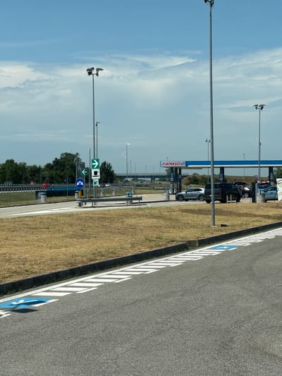 This is a photograph of a service station on a sunny day. In the background, a TAMOIL gas station is visible with several cars parked and refueling. The service station appears to be located in the city of Occimiano, Italy, indicated by the presence of Italian road signs. The foreground shows a portion of an asphalt road with white stripes and a blue handicapped parking symbol. The environment is characterized by dry, brown grass and scattered trees. The sky is partly cloudy.
