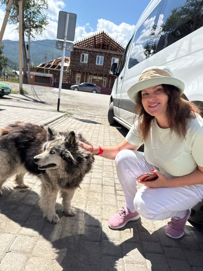 A woman with dark, wavy hair and a light-colored bucket hat is kneeling beside a large, shaggy dog with blue eyes on a paved outdoor area. She is wearing a light green t-shirt with "TRUSSARDI" embossed on it, white pants, and pink athletic shoes. The woman is smiling and holding a smartphone in her right hand while her left hand is touching the dog's head. The dog is looking to the left, with its mouth slightly open. The background shows a white van, a wooden building with exposed roof beams, other buildings, a green car, mountains in the distance, and a clear blue sky with scattered clouds. The scene appears to be in Onguday, Russia, given the architectural style of the buildings and the surrounding natural landscape.