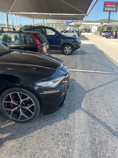 The image depicts a parking area under a large, grey fabric canopy, likely a car shelter at a service station or commercial establishment. In the foreground, the front section of a black car is visible, focusing on its wheel, tire, and a portion of the hood and headlight. The wheel is a multi-spoke alloy with red brake calipers. Behind the black car, a dark blue SUV is parked, followed by a black station wagon with a roof rack. Further in the background, other vehicles are parked in rows. A sign for "GOODY'S BURGER HOUSE" and "FLOCAFE" is prominently displayed on a tall pole to the right. The ground is asphalt. The context of Municipal Unit of Iera Poli Messologhiou, Greece, suggests this is a public parking area, possibly associated with a gas station and eatery, given the visible signage. The scene is outdoors on a sunny day, with shadows cast by the canopy and possibly by objects outside the frame. There are no people clearly visible in close proximity to the cars in the foreground, though a person can be seen walking near the vehicles in the mid-ground. The environment appears to be a commercial zone within a town or city.