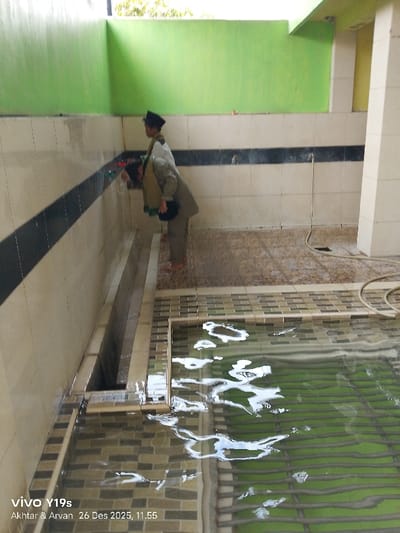 Two young boys in Islamic attire perform ritual ablutions at a tiled trough with faucets in a green and white tiled room. One boy leans over, drinking from a tap, while the other stands by. Water overflows onto a tiled floor and into a pool with a green slatted bottom. A hose lies coiled nearby. The scene appears to be a public or communal washing area.