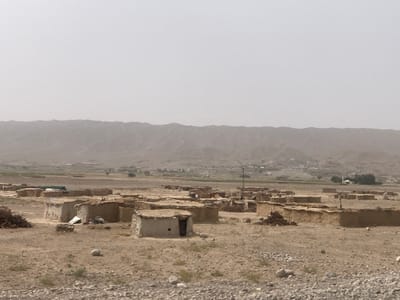 Dusty landscape with a village of small, mud-brick structures. Most buildings appear simple and square, with thatched roofs. In the background are barren hills under a hazy, overcast sky. The foreground is a rocky, arid plain with sparse vegetation. The overall impression is one of a remote, rural, and possibly impoverished setting.