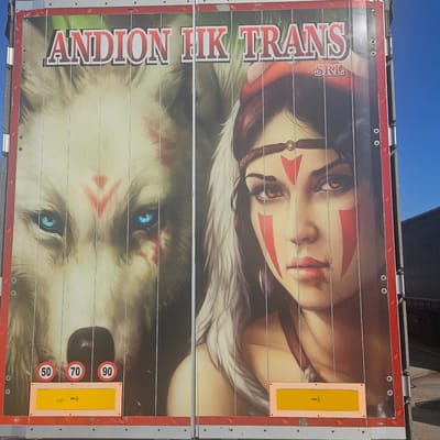The image shows the side of a truck trailer in Obrežje, Slovenia. The trailer features a large advertisement with a portrait of a woman with tribal face paint above a wolf with piercing blue eyes. The name "ANDION FIK TRANS SRL" is vertically printed on the left side of the advertisement. Two reflective yellow panels are on the right side, as well as speed limit signs (50, 70, 90). The trailer is metallic with a red frame and small silhouettes of elephants. The sky is visible above the trailer.