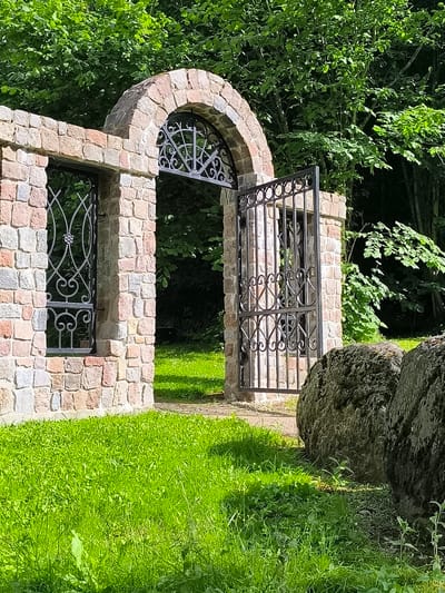 This is a photograph of an arched stone gateway with an open wrought-iron gate, located in the town of Mosėdis, Lithuania. The gateway is constructed from rough-hewn, multi-colored stones, and features a decorative wrought-iron panel within the arch. To the left of the gateway, a section of stone wall with a window protected by a similar wrought-iron grate is visible. The ground in front of the gateway is covered in lush green grass, and two large, moss-covered rocks are positioned in the foreground to the right. Behind the gateway and wall, dense green foliage from trees forms a natural backdrop. The sunlight creates a dappled effect on the grass and stone.