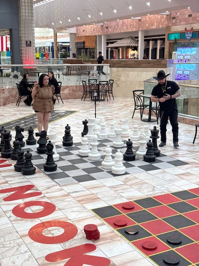 In El Paso, United States of America, within a well-lit indoor shopping mall, a large outdoor chess set is positioned on a checkered floor. The chessboard is approximately 30x30 feet, with alternating white and light brown marbled tiles. Several giant chess pieces, both black and white, are arranged on the board. In the foreground, to the right of the chessboard, a checkerboard pattern with red and black squares is visible, with red and black checkers placed on some of the squares.
In the midground, standing to the right of the chess set and facing it, is a man wearing a black cowboy hat, a black t-shirt, dark pants, and black boots. He has tattoos on his arms and is wearing a gold necklace. His hands are clasped in front of him. To the left of the chess set, a woman wearing a leopard-print dress and dark shorts is standing. Behind her, two other women in black attire are seated at a table.
In the background, the mall environment is visible with various shops, storefronts, and decorative lighting. Some tables and chairs for diners are also present. A security guard in a dark uniform is seen in the distance near a stairway. The scene suggests a recreational activity or a public gathering within the mall.