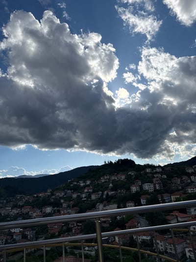 The image shows a hillside view from Momchilovtsi, Bulgaria. Houses are densely packed along the hillside, framed by a metal railing in the foreground. The sky is partly cloudy with some gaps where the sun shines through.