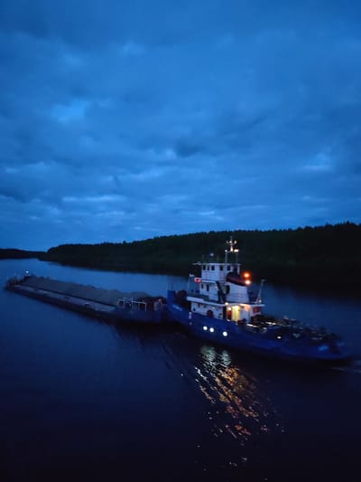 The image depicts a barge being towed by a tugboat on a waterway, likely a river or canal, in Alexandrovskoye, Russia. The tugboat is brightly lit with multiple lights, reflecting on the water surface. The barge is loaded with a dark, granular material, possibly gravel or sand. The scene appears to be at dusk or night, with a dark, cloudy sky overhead and wooded banks lining the waterway.