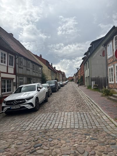 This is a photograph taken in Plau am See, Germany, depicting a street scene. The street is paved with cobblestones, with a slightly raised brick sidewalk on the right. The perspective is from street level, looking up a gently sloping street lined with buildings on both sides. The sky is overcast with dramatic clouds.
On the left side of the street, a white Mercedes-Benz SUV is parked in the foreground. Behind it, a row of cars is parked along the curb. The buildings on the left are a mix of architectural styles, with some featuring traditional half-timbered construction and red tiled roofs.
On the right side, the buildings are also varied, with one prominent structure made of red brick with white-framed windows. Further down the street, other buildings of varying heights and colors line the road. A wooden fence runs along the side of the property on the right. There is a metal street lamp and some greenery visible. The overall impression is of an old European town street on a cloudy day.