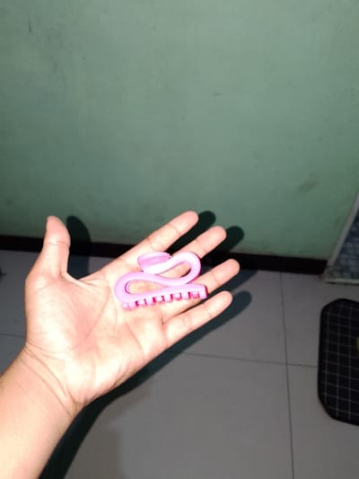 A close-up shot shows a person's left hand and forearm holding a pink hair clip. The hair clip is curved and has small teeth along its inner edge. The hand is open, with the palm facing upwards, and the hair clip is positioned across the palm. The background consists of a plain light green wall and a tiled floor. Shadows from the hand and the hair clip are visible on the wall and floor. A black and white checkered object is partially visible on the floor to the right. The scene appears to be indoors.
