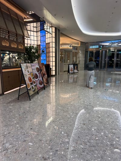 The image depicts an interior space, likely a shopping mall, with a polished stone floor. In the center-right, a person wearing a denim jacket and light-colored trousers is walking away from the camera. A large restaurant is on the left with a sign in Chinese characters hanging overhead. There are menu boards on stands in front of the restaurant showing pictures of food. This location is in the Jianye District of China.